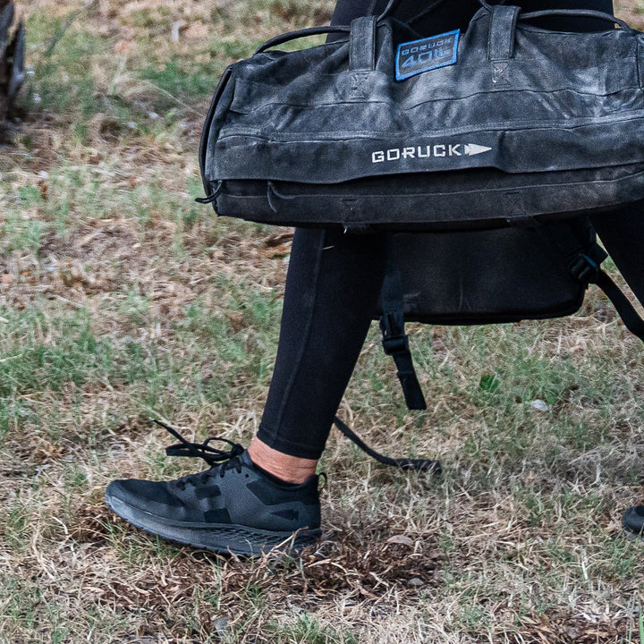 A person strides across the grass, wearing black leggings and sleek running shoes with a Gradient Density EVA midsole for comfort, while carrying a GORUCK Rough Runner - Blackout bag. The road-to-trail outsole of the shoes effortlessly adapts to changing terrains.