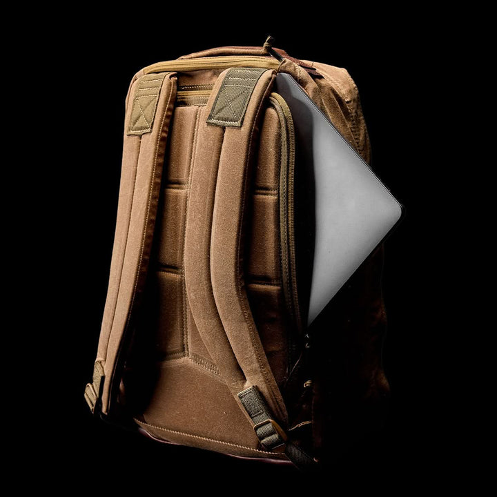 The GORUCK GR1 USA - Heritage is a durable brown waxed canvas backpack with a visible laptop in the side pocket, shown against a black background.