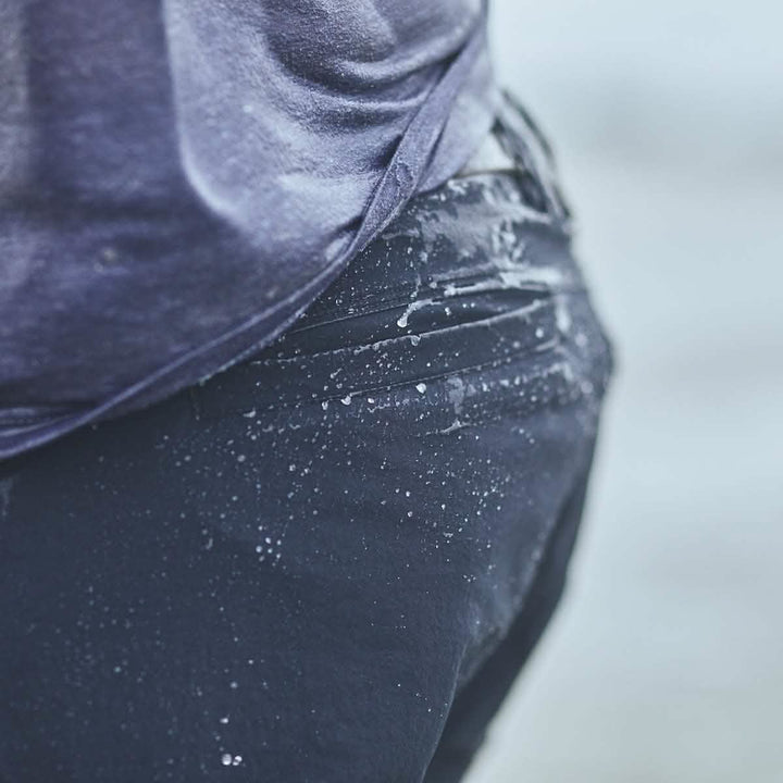 Close-up of a person wearing wet dark Men’s Challenge Shorts from GORUCK, made with Lightweight ToughDry® fabric, featuring water droplets glistening on the material. The shorts are equipped with practical cargo pockets and a diamond gusset for comfort, suggesting they are near a body of water outdoors.