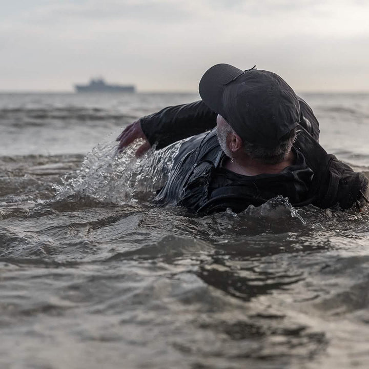 A person wearing a GORUCK Performance TAC Hat made from ToUGHTDRY® fabric floats effortlessly in the sea, reaching towards a distant ship on the horizon.