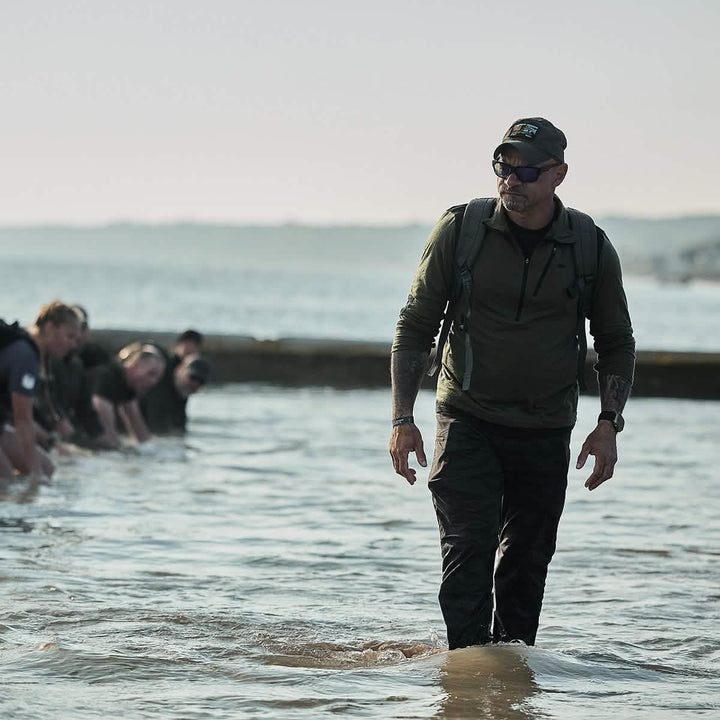 A man in tactical gear, wearing the Performance TAC Hat made by GORUCK with TOUGHDRY® fabric, walks through water as others crawl behind him.