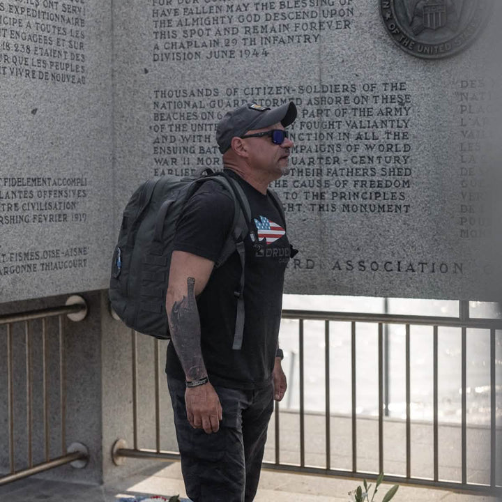 A man in sunglasses and a ToughDry® fabric backpack stands reading a wall inscribed with text, while his GORUCK Performance TAC Hat - TOUGHDRY shields him from the sun.