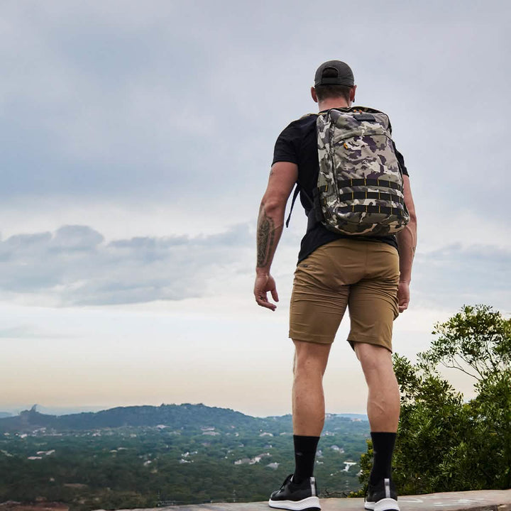 A person wearing a camouflage backpack and black cap stands on a hilltop, taking in the scenic landscape of hills under a cloudy sky. They are dressed in a black shirt and GORUCK's Men’s Challenge Shorts made from lightweight ToughDry® fabric, and they complete their look with black shoes.