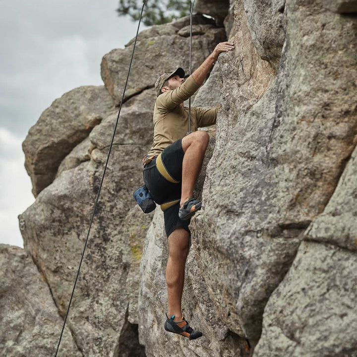 A person sporting the Men’s Challenge Shorts by GORUCK, featuring a cap and cargo pockets, is rock climbing on a rocky cliff. They are equipped with a harness and climbing shoes, focused and reaching for a handhold against a cloudy sky backdrop.