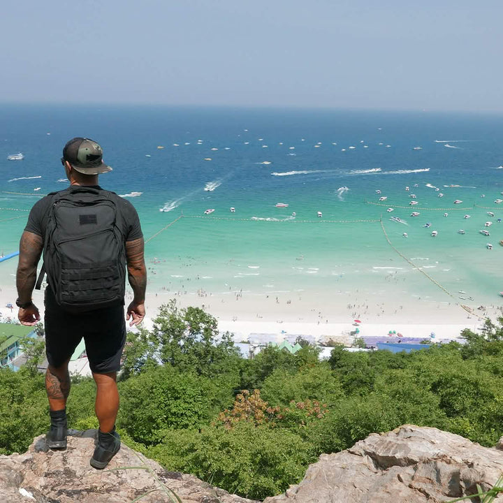 A person wearing a backpack and GORUCK Men’s Challenge Shorts in Lightweight ToughDry® fabric stands on a rocky ledge overlooking a beach with turquoise water. Boats and people can be seen in the ocean and along the sandy shore under a clear blue sky.