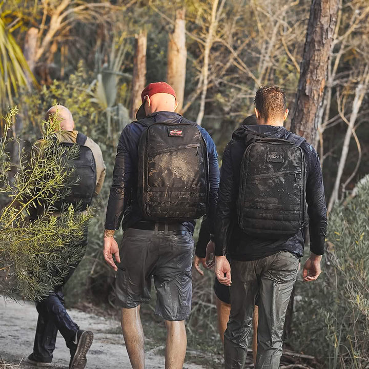 Three individuals are hiking through a muddy trail, dressed in outdoor gear equipped with cargo pockets and backpacks. The forested area, rich with trees and shrubs, surrounds them as they proceed away from the camera. Despite being soaked from the rain, their GORUCK Men’s Challenge Shorts made from Lightweight ToughDry® material are perfectly suited for wet conditions.