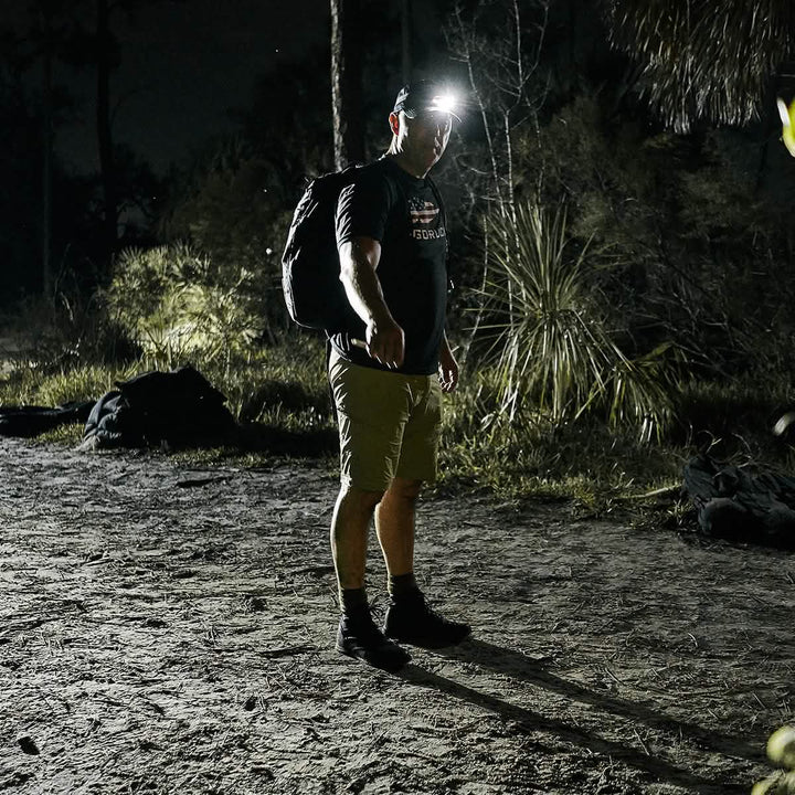 In a dimly lit outdoor setting, a man is highlighted by the glow of a headlamp. He's clad in a black shirt and khaki GORUCK Men’s Challenge Shorts - Lightweight ToughDry®, complete with cargo pockets, while carrying a backpack. The scene is enhanced by the presence of trees and bushes, contributing to the nighttime ambiance.