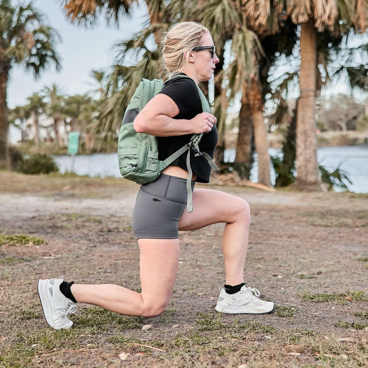 A person wearing the Women’s Squat Shorts - ToughFlex by GORUCK is doing lunges outside near the trees and water, enjoying a perfect blend of nature and comfort.