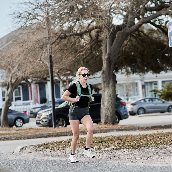 A person jogs on a sidewalk, wearing sunglasses and a backpack. They sport GORUCK's Women’s Squat Shorts made with ToughFlex Fabric, ensuring comfort amidst the trees and parked cars lining the path.