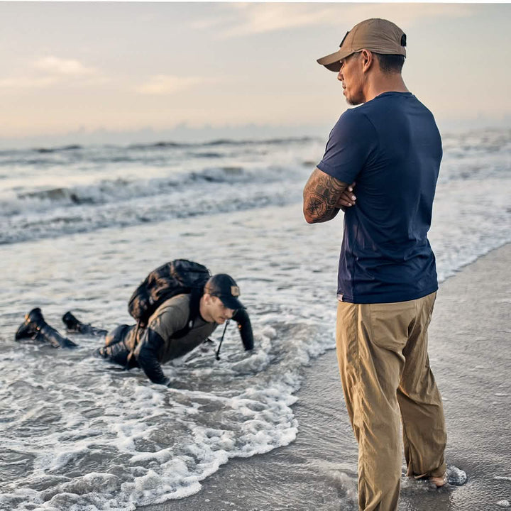 Two men on a beach; one stands with arms crossed, wearing a GORUCK Performance TAC Hat made of TOUGHDRY fabric, while the other crawls through the surf with a VELCRO® strapped backpack.