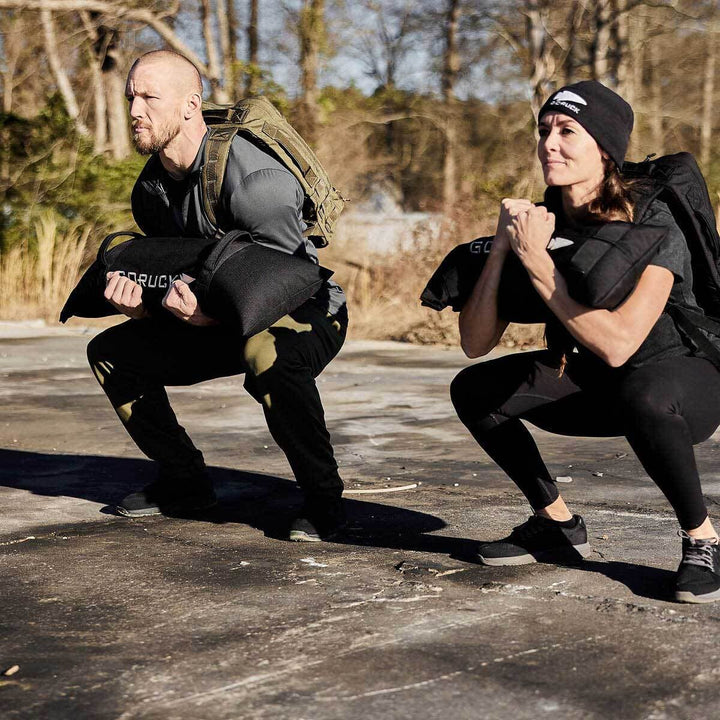 Two individuals showcasing their commitment to fitness outdoors, squatting with GORUCK's Simple Training Sandbags, dressed in athletic clothing and beanies.