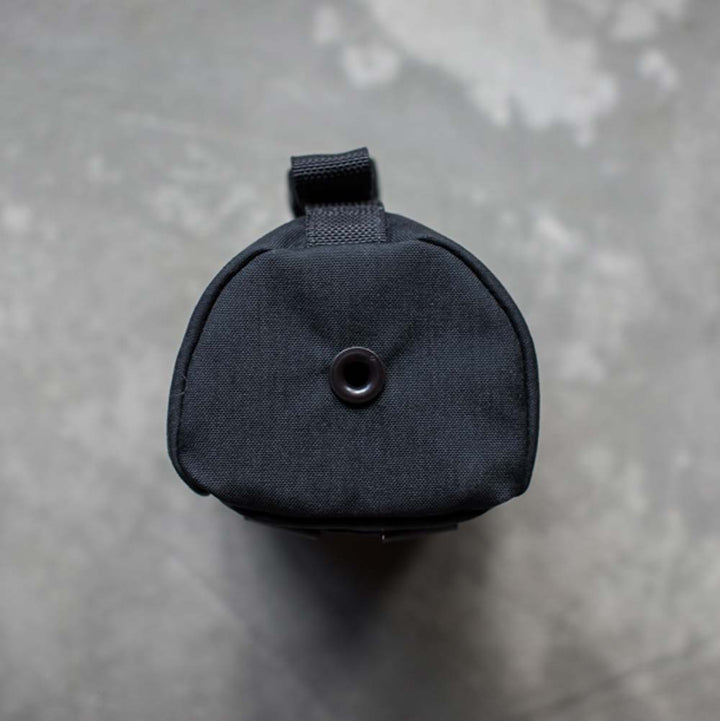 Discover the GORUCK Full Panel Water Bottle Pocket, a black cylindrical fabric bag with a strap, known for its sleek design that suits the minimalist traveler. Pictured from above against a textured gray background, it features an ideal pocket for your Nalgene-style water bottle, ensuring easy hydration on the go.