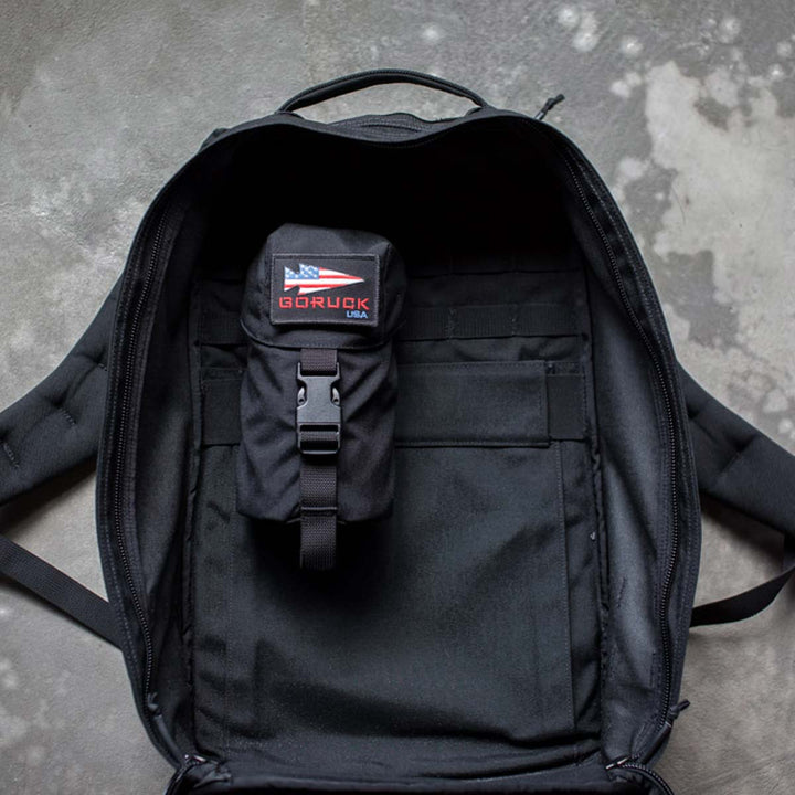 Open the GORUCK Full Panel Water Bottle Pocket, a black backpack equipped with a small black pouch inside, showcasing a USA flag and GORUCK branding. Designed with a MOLLE attachment system, it is ideal for securing gear while traveling. The bag also features a handy pocket to securely hold your Nalgene-style water bottle.