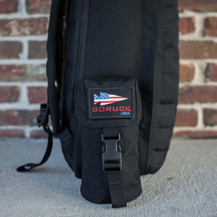 GORUCK's Full Panel Water Bottle Pocket, a black backpack featuring an American flag patch and the "GORUCK USA" label, rests against a brick wall. It includes a pocket designed for your Nalgene-style water bottle and incorporates the MOLLE attachment system for added versatility and rugged style.