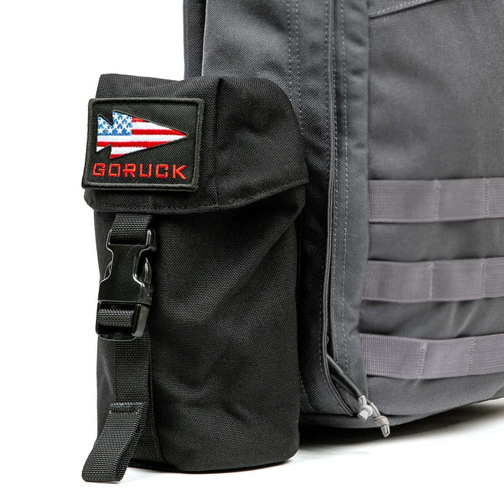 Close-up of a gray backpack with a black side pouch showcasing a "GORUCK" patch alongside an American flag design. Featuring the GORUCK Full Panel Water Bottle Pocket, this backpack utilizes a MOLLE attachment system to securely hold a Nalgene-style water bottle, keeping you hydrated during your adventures.