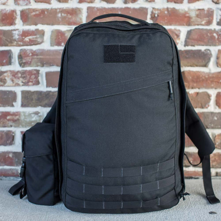 A GORUCK Full Panel Water Bottle Pocket in black, featuring a detachable side pouch and MOLLE attachment system, is set against a brick wall.