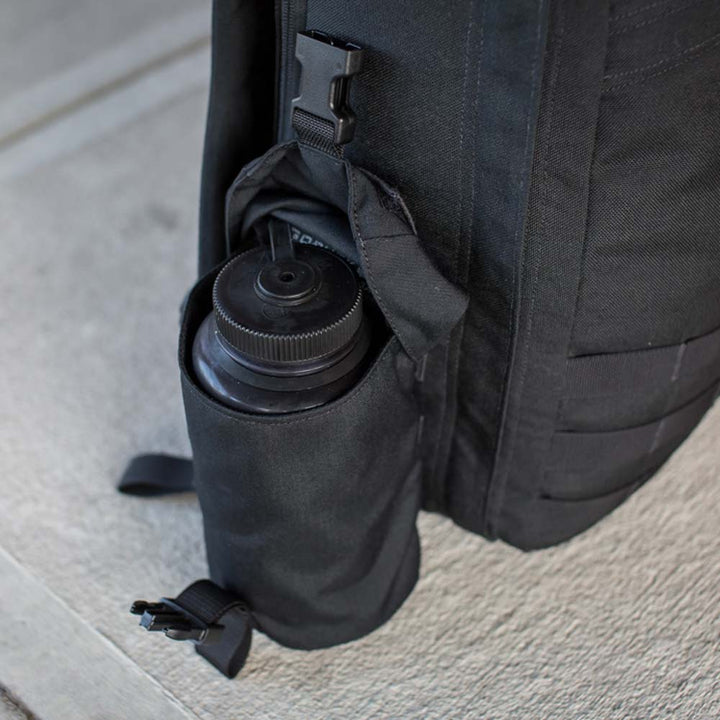 A GORUCK backpack with a Full Panel Water Bottle Pocket holds a Nalgene-style water bottle snugly, resting on a concrete surface.