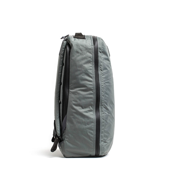 The GORUCK GR1 USA - X-PAC backpack stands upright on a white background, showcasing a vertical AquaGuard zipper and black shoulder straps. Its durable X-PAC construction offers both reliability and style for any adventure.