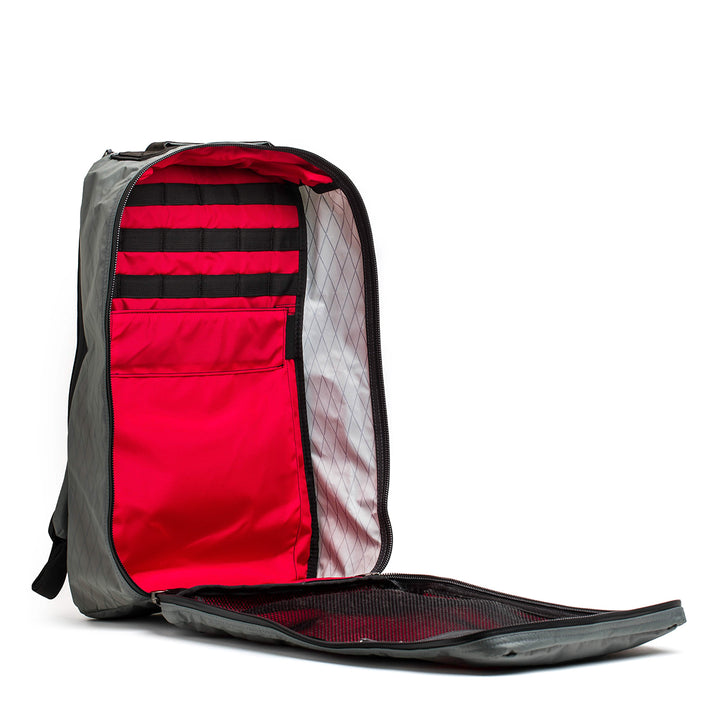 The GORUCK GR1 USA - X-PAC in grey, with AquaGuard zippers, is shown completely unzipped to display red inner compartments and black pockets.