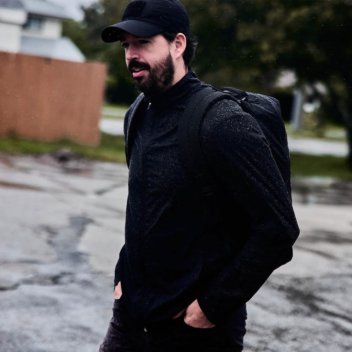 A man wearing a black cap and jacket walks outside on a rainy day with the GORUCK GR1 USA - X-PAC backpack, featuring AquaGuard zippers to keep his belongings safe and dry.