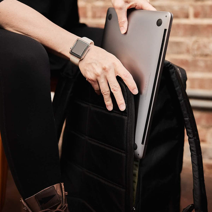 A person carefully places their laptop into a sleek GR1 USA - X-PAC backpack by GORUCK, equipped with AquaGuard zippers, while checking their smartwatch.