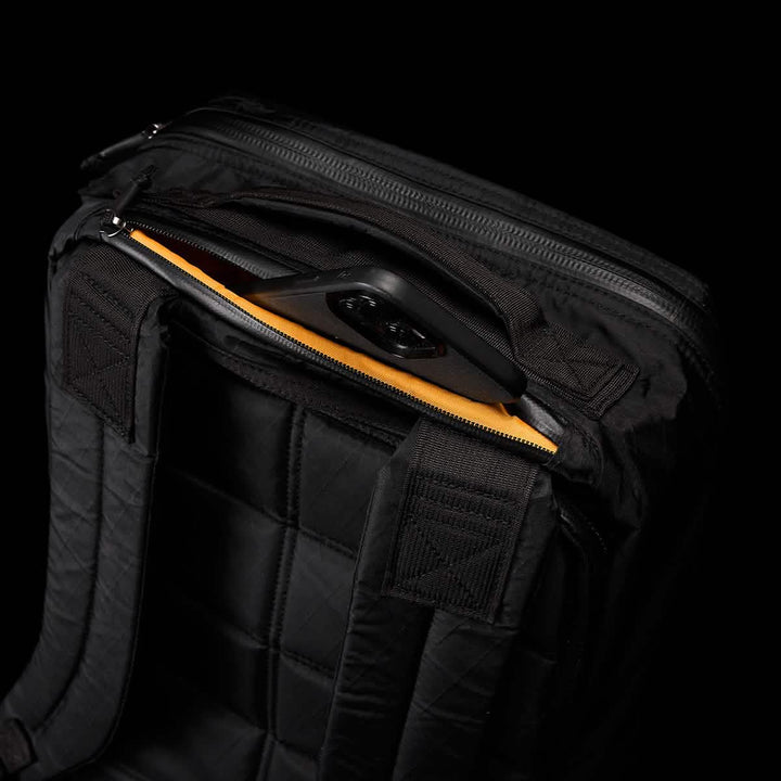 The GORUCK GR1 USA - X-PAC black backpack showcases AquaGuard zippers and a quilted design pocket, partially open to reveal an orange lining and a device. It's crafted with water-resistant materials for all-weather protection.