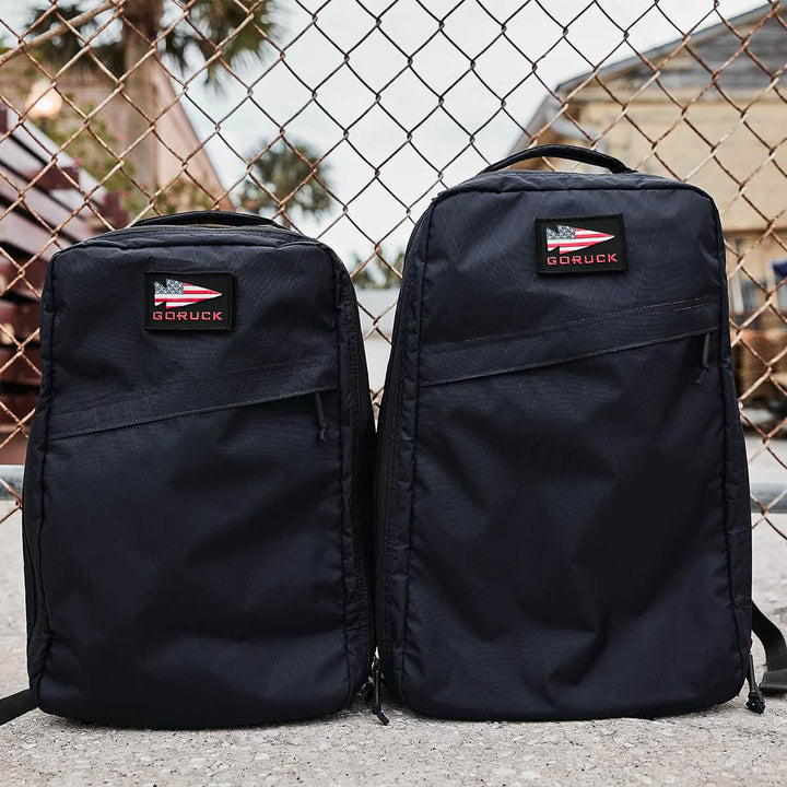 Two GORUCK GR1 USA - X-PAC black backpacks, featuring AquaGuard zippers and logo patches, are placed in front of a chain-link fence outdoors.