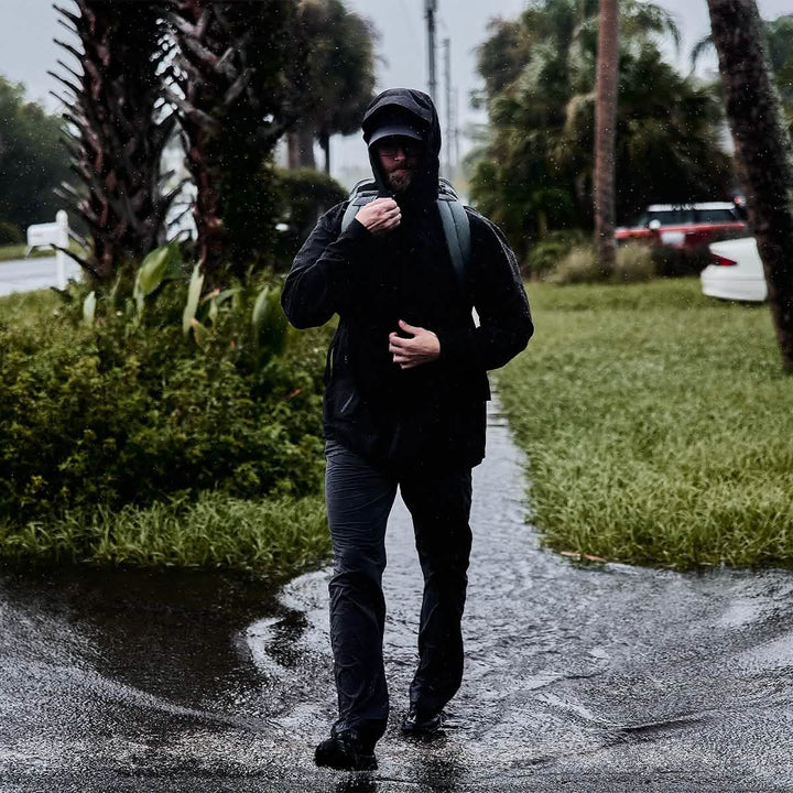 Wearing GORUCK's GR1 USA - X-PAC with AquaGuard zippers, an individual strolls along a puddle-covered path, surrounded by lush greenery, as cars loom in the distance.