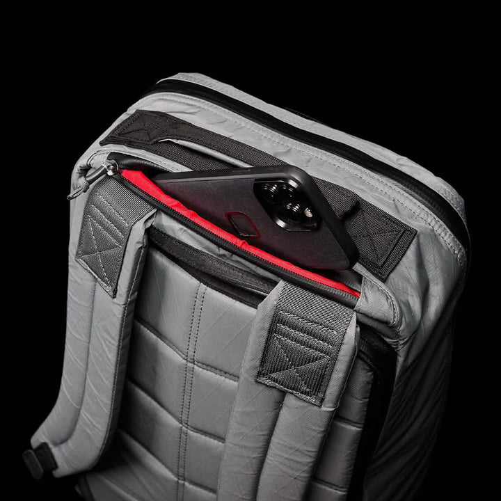 The GORUCK GR1 USA - X-PAC backpack in grey has AquaGuard zippers, with a red-lined pocket partially unzipped to reveal a smartphone in a black case.