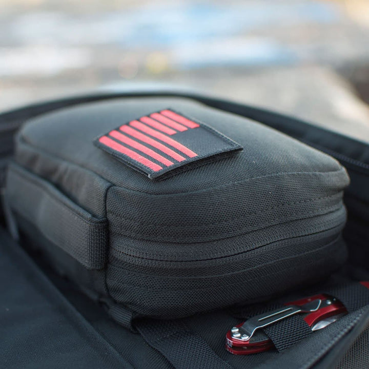 A GR1 Field Pocket by GORUCK, featuring a sleek black design with red stripes, is strategically placed in an open backpack and serves as an electronics organizer alongside a multi-tool.