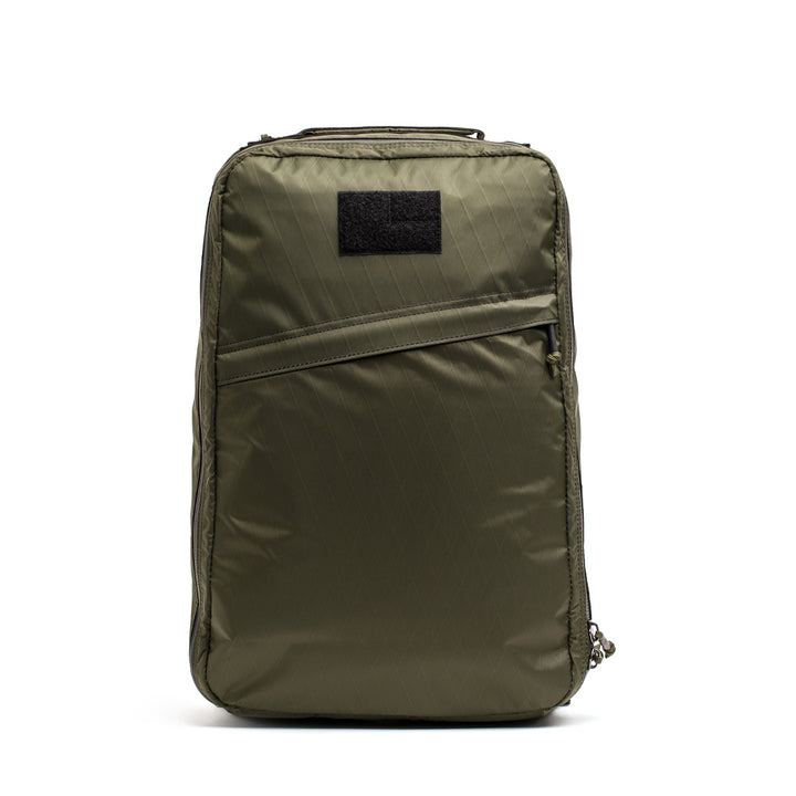 The GORUCK GR1 USA - X-PAC backpack in olive green boasts a durable design with a rectangular patch, diagonal front pocket, and AquaGuard zippers on the side and top for waterproof protection.