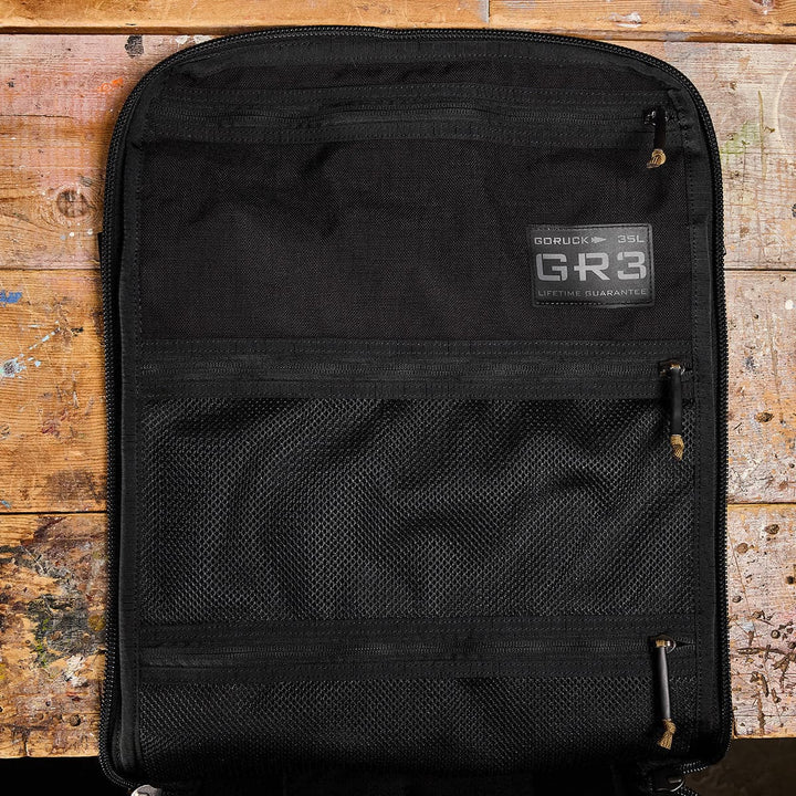 The GR3 - Cordura by GORUCK, a durable black backpack designed for travel, rests flat on a rustic wooden surface. Opened to display two mesh compartments and the GR3® 35L label, it features water-resistant properties that accentuate the visible paint splatters and scratches beneath.