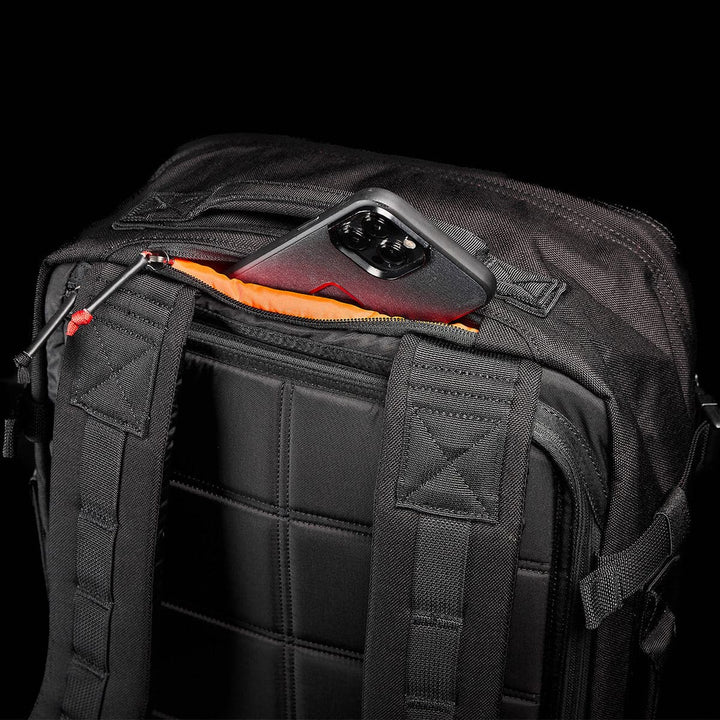 A GR3 - Cordura carry-on rucksack from GORUCK rests on its side, with a smartphone encased in metallic peeking out of an open pocket. This sleek black backpack showcases water-resistant material, padded straps, and a quilted GR3® design on the back.