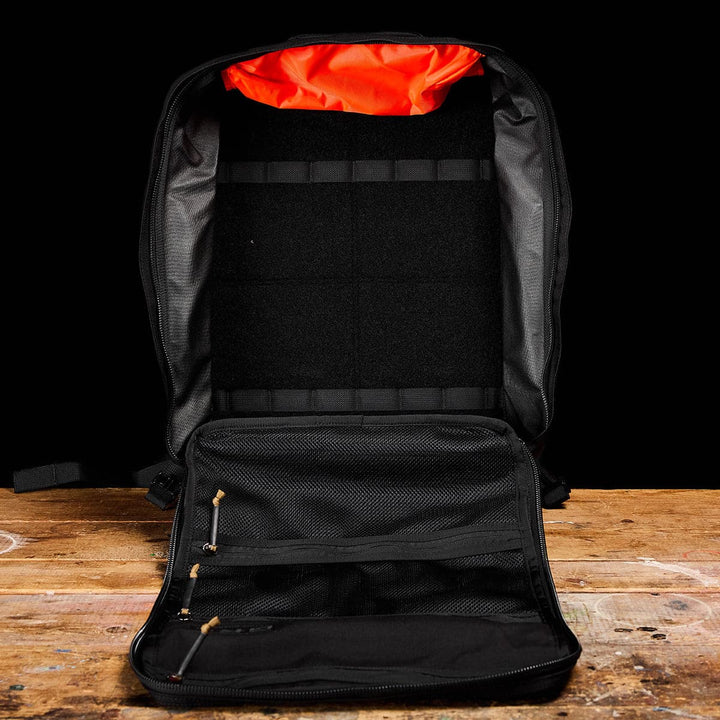 The GR3 - Cordura by GORUCK, a black backpack ideal as a carry-on rucksack, is shown open on a wooden surface. Its interior features a bright orange lining and several pockets for organization. The spacious main compartment is partially padded, making it an excellent choice for travel with its water-resistant design.