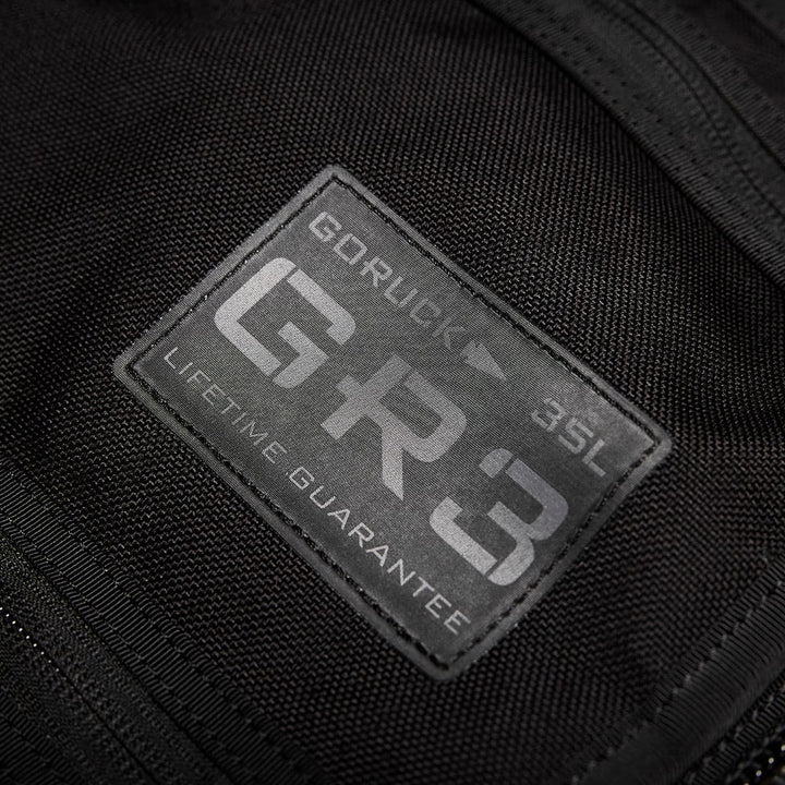 Close-up of a black fabric patch on a carry-on rucksack displaying the text: GORUCK - GR3® - Cordura LIFETIME GUARANTEE. The rugged, water-resistant patch is stitched onto the fabric, highlighting durability and quality.