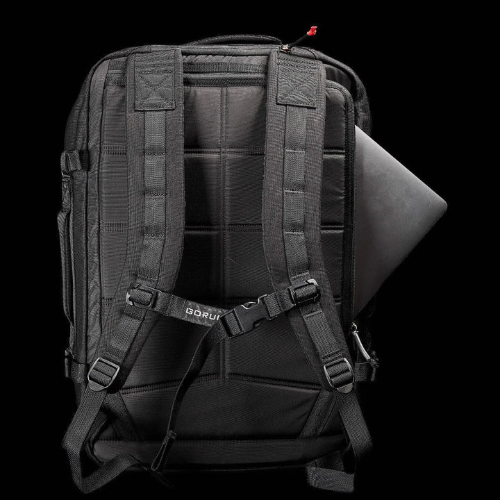 The GR3 - Cordura by GORUCK is a black carry-on rucksack featuring adjustable padded straps and a visible brand tag, complete with a square pattern on the back. A corner of a laptop can be seen tucked away in the backpack's water-resistant compartment against a solid black background.