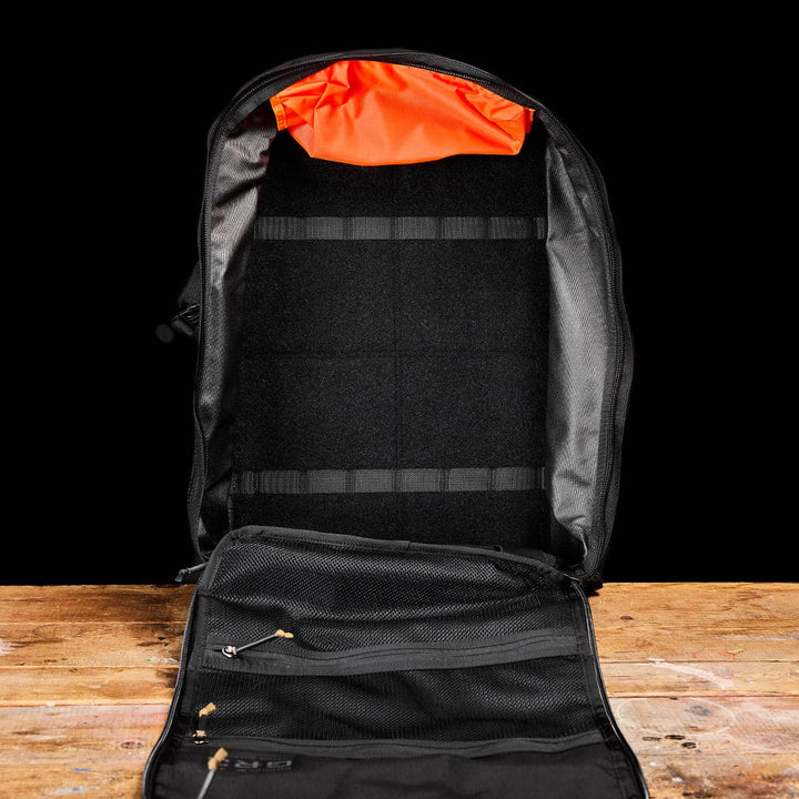 Place the open GR3 - Cordura backpack by GORUCK on a wooden table. It showcases a bright orange lining at the top, complemented by several black compartments and mesh pockets inside, emphasizing its convenience as a carry-on rucksack. Made with water-resistant materials, it's ideal against a solid black backdrop.