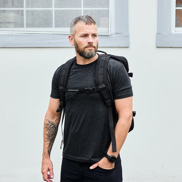 Outdoors, a bearded man with short hair is wearing a GORUCK GR3 - Cordura, a water-resistant black carry-on rucksack, paired with a dark T-shirt. He has a tattoo on his right arm and gazes to the side. In the background, there's a light-colored building with a window, enhancing his adventurous style.