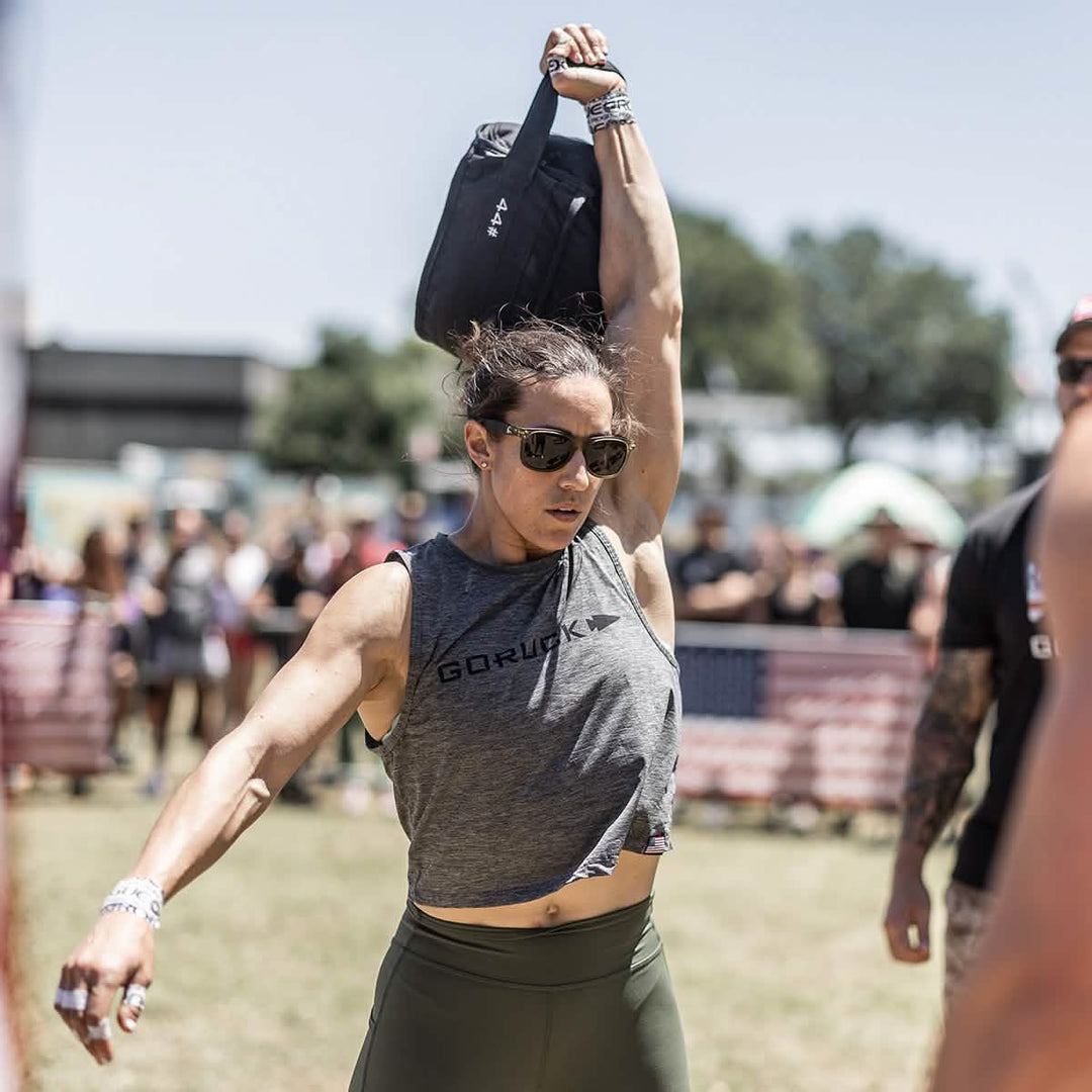 During an outdoor event, a person in sunglasses and athletic wear lifts a weight overhead, highlighting the durability of their GORUCK Women’s Performance Tank - ToughMesh. In the background, a crowd and American flag banners are visible under a sunny sky.