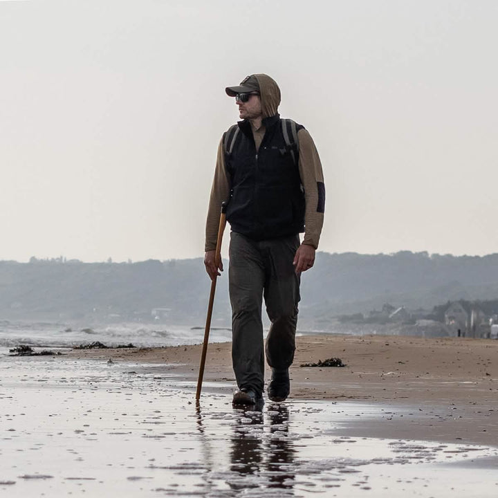 A person strolls along the beach with a stick, wearing a GORUCK Performance TAC Hat made of ToughDry® fabric and sunglasses, under a cloudy sky.