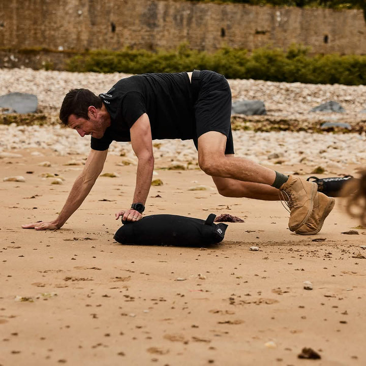 A man exercises effortlessly on a sandy beach, balancing on a sandbag while wearing lightweight MACV-1 Hi-Speed boots by GORUCK.