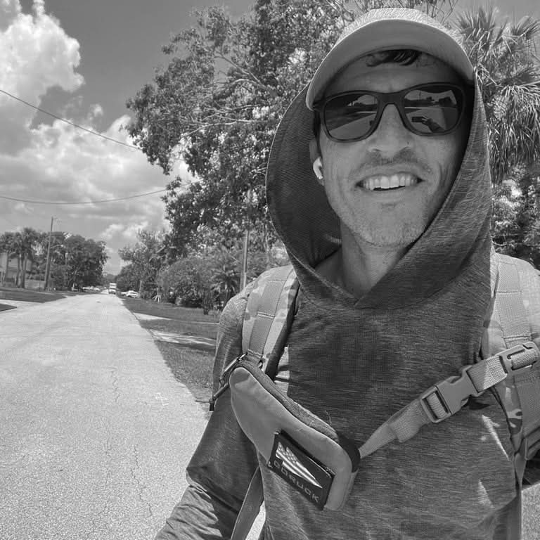 A person wearing sunglasses, a hat, and a hooded jacket smiles at the camera. They have a backpack and a sleek Sternum Pouch - Cordura from GORUCK. The background shows a sunny street lined with trees and a partly cloudy sky.