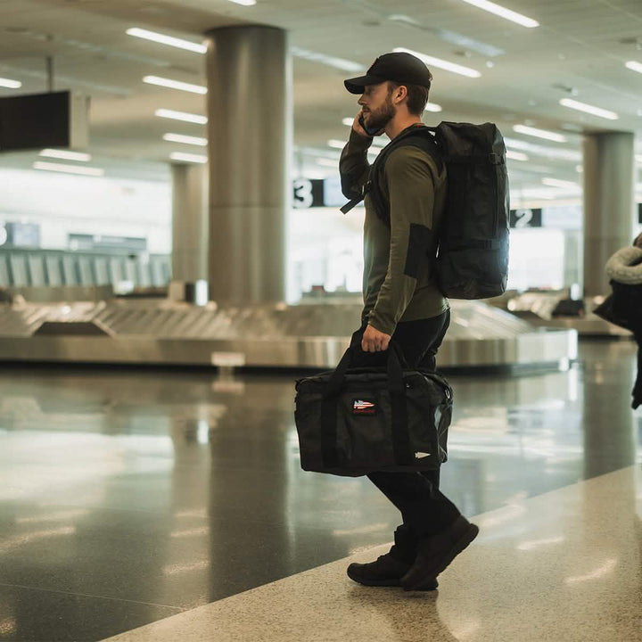 A man in a cap and casual attire strolls through an airport, carrying a large GORUCK GR3 - Dyneema backpack and a duffel bag. Crafted from durable Dyneema material, his gear guarantees resilience. Conversing on his phone, he effortlessly navigates past baggage claim carousels in the brightly lit terminal.