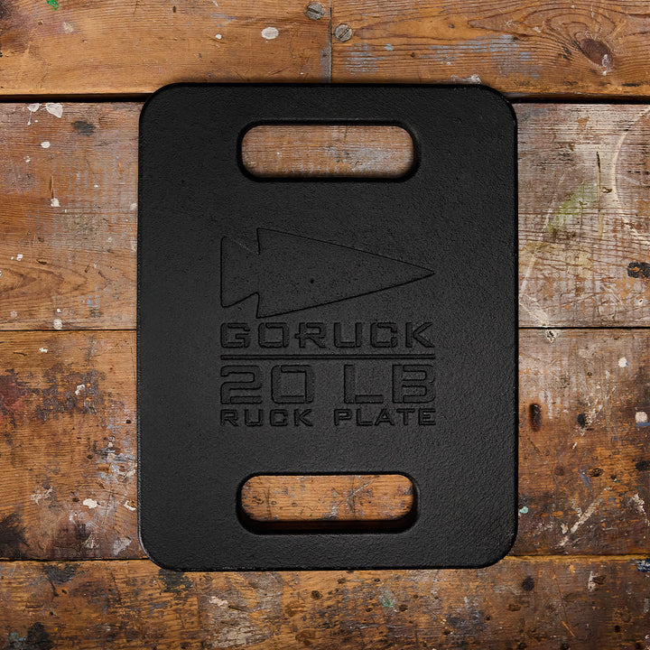 A sturdy 20 lb black GORUCK Ruck Plate with cutout handles sits on a worn wooden surface, perfect for your Rucker or Ruck Plate Carrier.