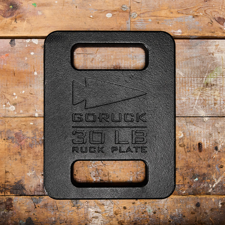 A GORUCK Ruck Plate in black, weighing 30 lbs and featuring handles, rests on a wooden surface. It's perfect for pairing with your Ruck Plate Carrier for intense workouts or with your reliable Rucker for exciting treks.