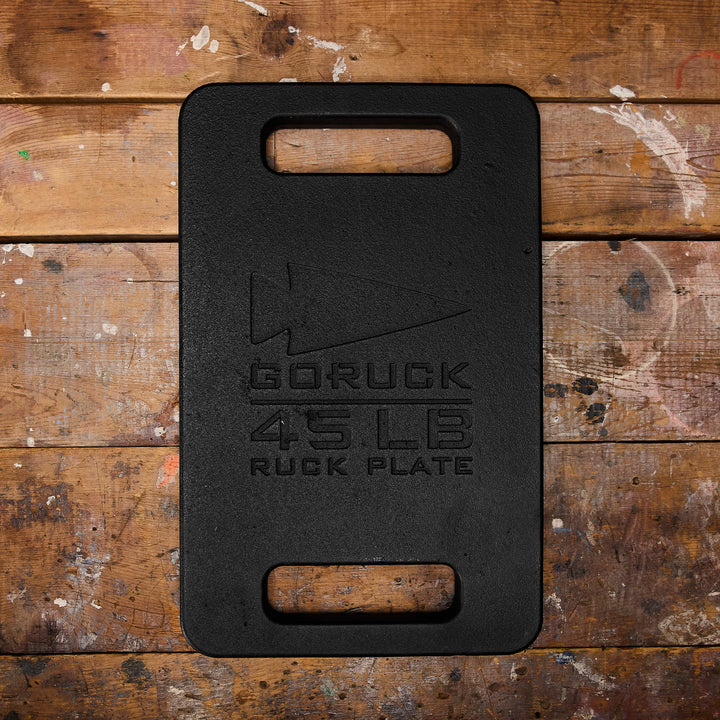 A black GORUCK Ruck Plate (45 LB) rests on a worn wooden surface, ready to fit seamlessly into your Ruck Plate Carrier.