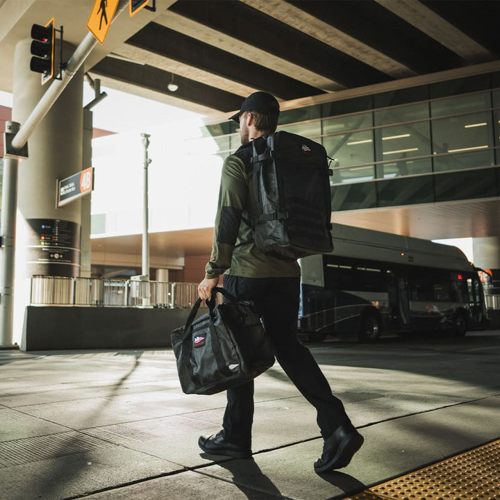 A person wearing a cap and dark clothing strides confidently across the street, making a statement with their MACV-1 Traveler boots from GORUCK. They carry a black backpack and another bag in an urban setting, framed by a bus and overhead road structures.