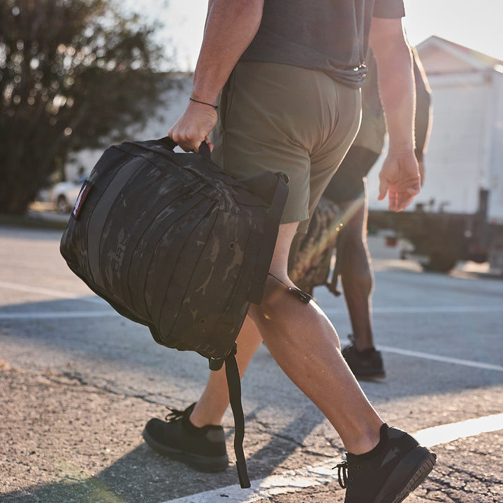 Dressed in athletic gear, a person walks through a sunny parking lot with the rugged GORUCK Rucker 4.0 rucksack, embodying the spirit of rucking.