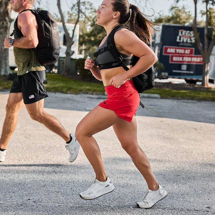 On a sunny day, two people are running outdoors wearing weighted vests and backpacks, dressed in squat-proof GORUCK Women's Training Shorts made from ToughStretch fabric for maximum comfort and durability.