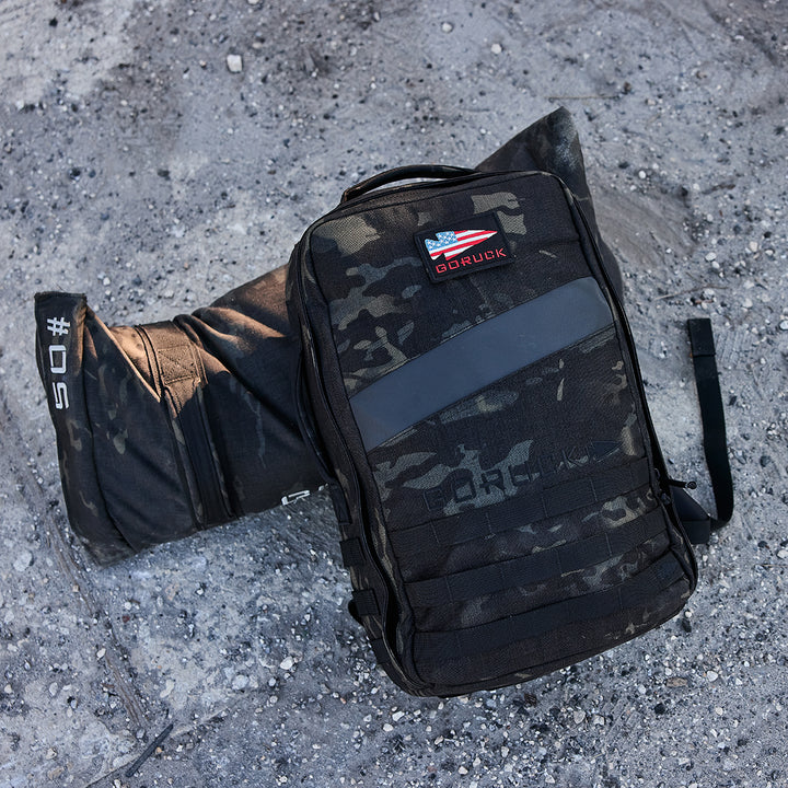 A black camo GORUCK Rucker 4.0 and a numbered sandbag rest on a rough concrete surface, ideal for your next rucking adventure.